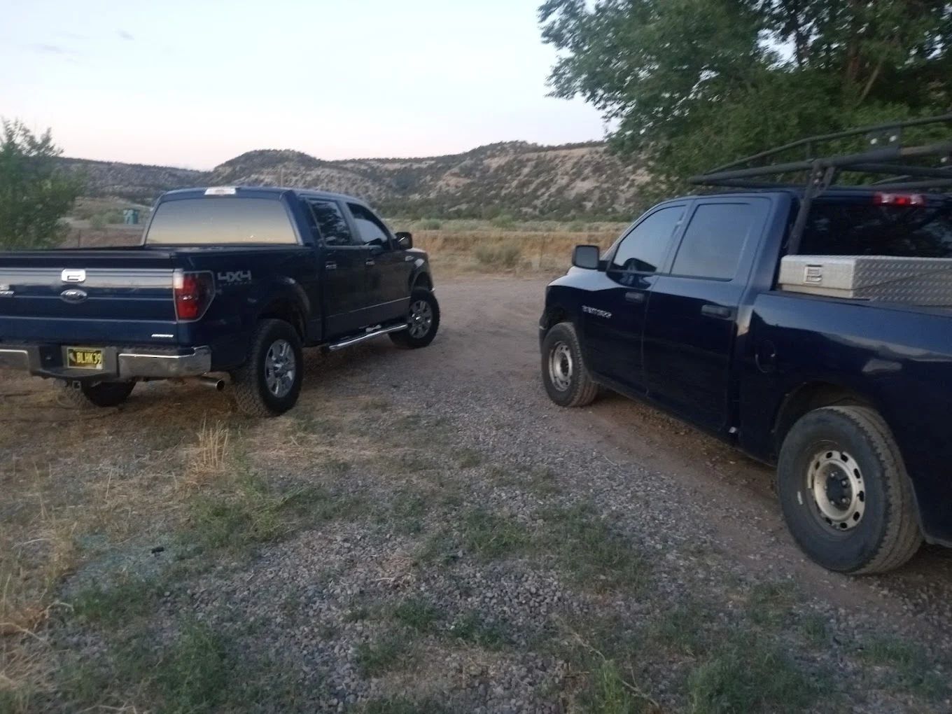 Two dark blue pickup trucks parked on gravel with desert hills in background
