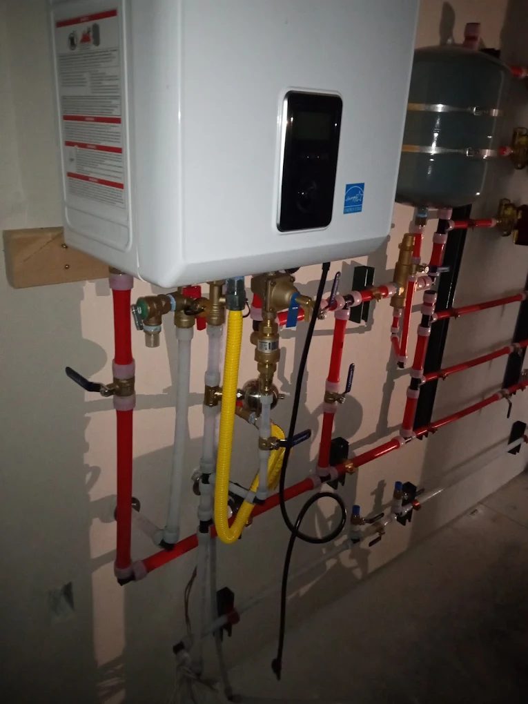 Tankless water heater with digital display mounted on wall above red and white PEX tubing manifold system with brass fittings and valves