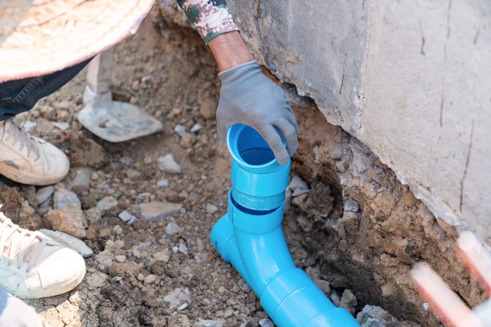 Worker in gray gloves installing blue PVC pipe in soil trench next to concrete foundation