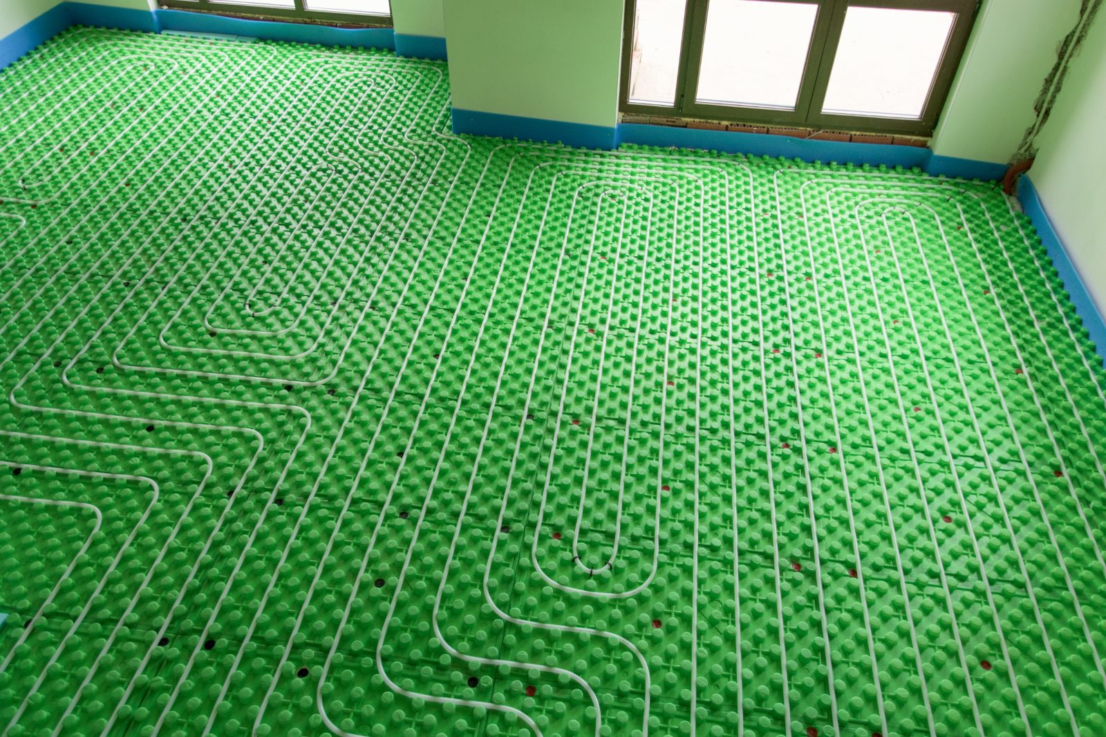 Radiant floor heating system with white tubing laid in serpentine pattern on green substrate in empty room