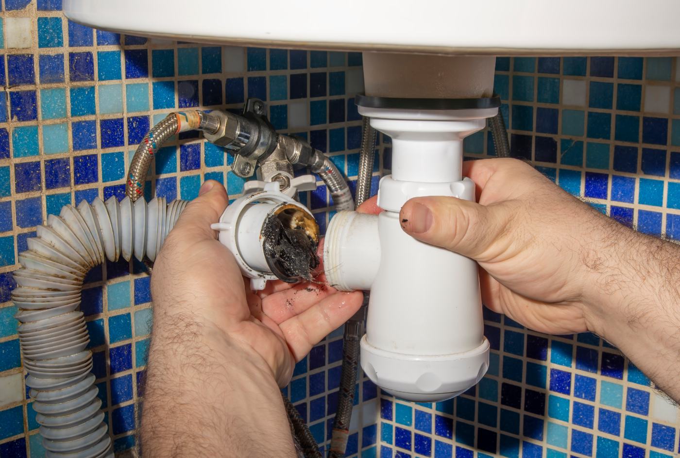 Plumber's hands removing clogged P-trap from sink drain against blue tile backsplash