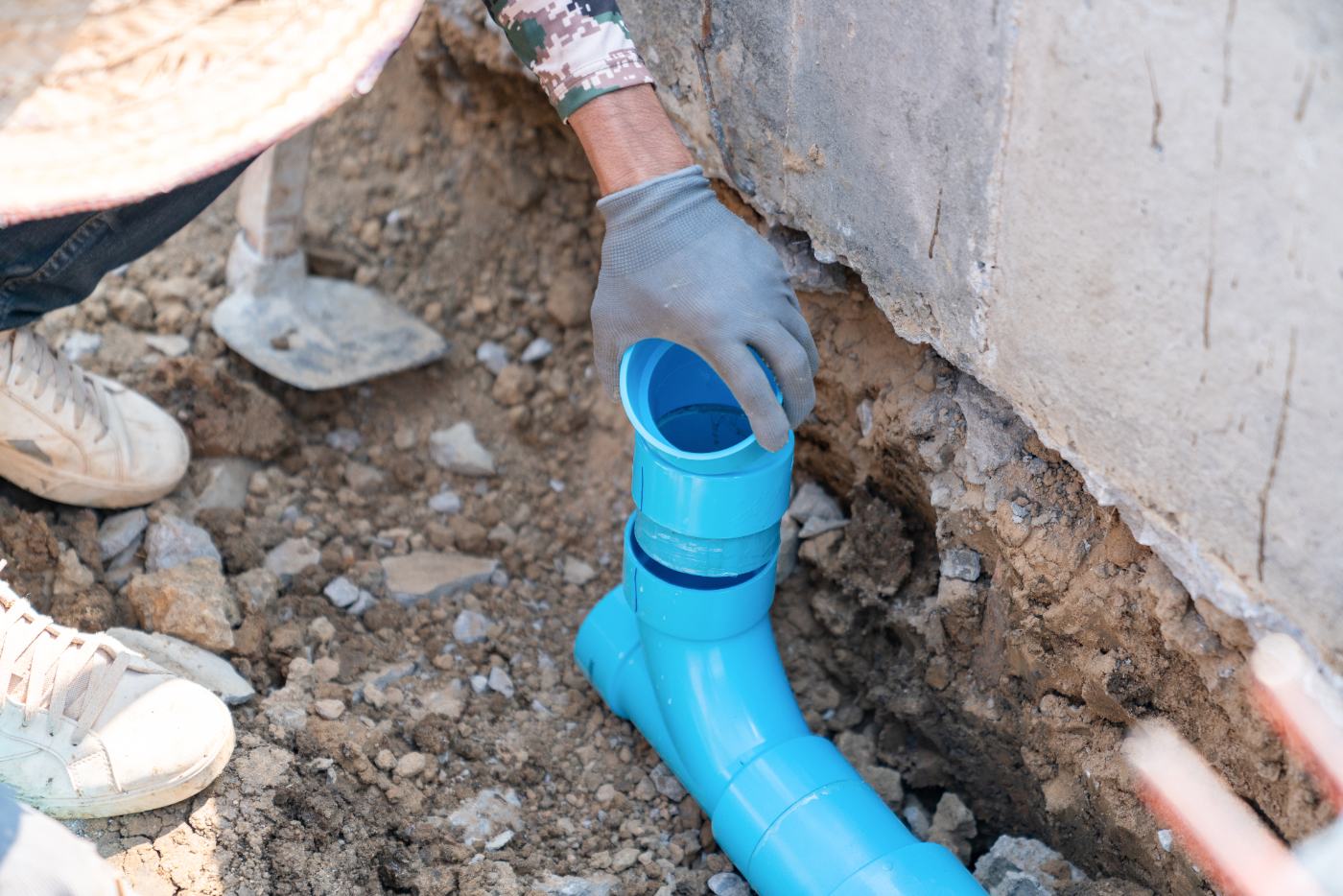 Worker in gray gloves installing blue PVC pipe in trench next to concrete foundation